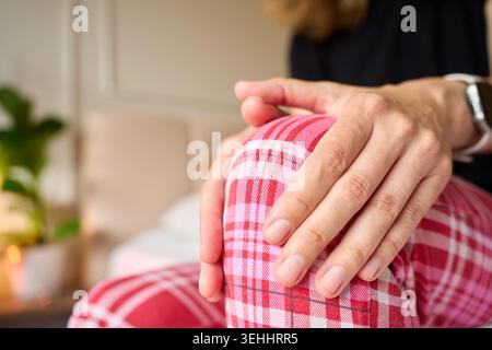 La donna soffre di dolore al ginocchio, massaggiando il ginocchio con entrambe le mani mentre è seduta sul letto. Concetto di mal di ginocchio, disagio articolare, problema di mobilità e casa Foto Stock