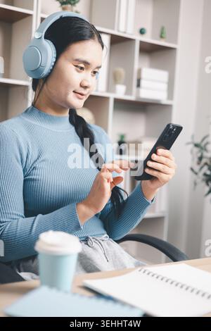 Giovane donna asiatica che indossa le cuffie e utilizza lo smartphone accanto al computer portatile alla scrivania. Apprendimento mobile, classe online, studio remoto, istruzione digitale A. Foto Stock