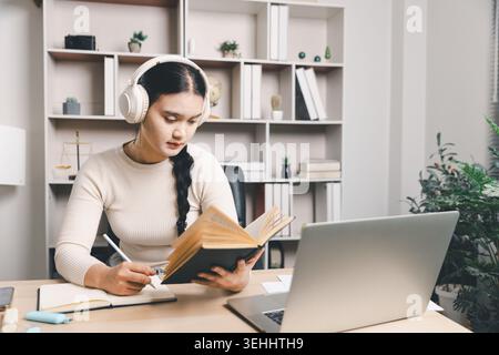Giovane donna asiatica che studia a casa con le cuffie, legge un libro, prende appunti e utilizza un computer portatile alla scrivania. Apprendimento online, formazione mirata, remoto Foto Stock