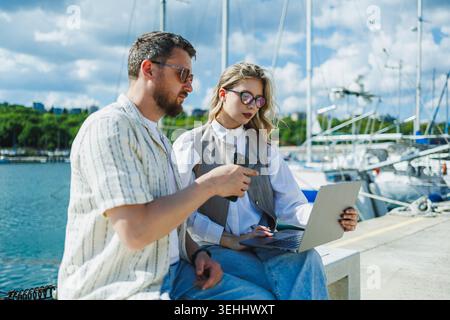 Due persone sono sedute vicino all'acqua al porticciolo. Stanno lavorando su un notebook e discutendo qualcosa. Le barche si trovano nelle vicinanze sotto un cielo limpido con clou Foto Stock