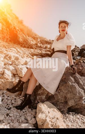 Donna di moda costiera elegante signora in abito bianco seduta sulle rocce durante l'ora d'oro al tramonto sul mare Foto Stock