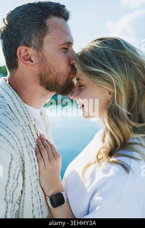 Una coppia si trova vicino all'acqua, godendosi una giornata di sole. L'uomo bacia la fronte della donna mentre lei si appoggia a lui, mostrando la loro connessione e io Foto Stock