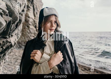 Donna ushanka Coast giovane donna che indossa una giacca di pelle e un cappello invernale si staglia sulla costa rocciosa del mare in un'atmosfera penosa all'aperto Foto Stock
