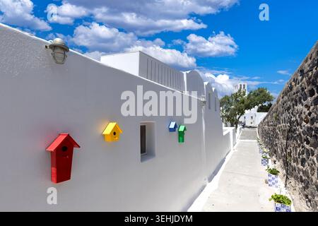Vista delle colorate case per uccelli montate su un muro bianco lungo una stretta strada sotto un cielo blu con nuvole a Fira, Grecia. Foto Stock