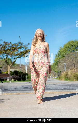 Donna matura felice che cammina a piedi nudi nel parco cittadino in abito floreale Foto Stock