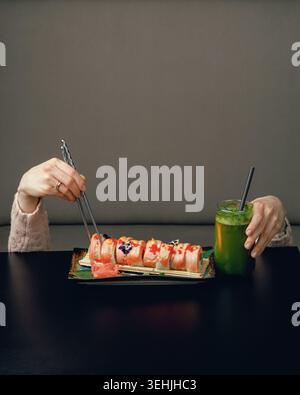 Una ragazza sotto un tavolo con le bacchette sul tavolo mangia sushi e beve un cocktail. creatività, pubblicità, spazio per il testo Foto Stock