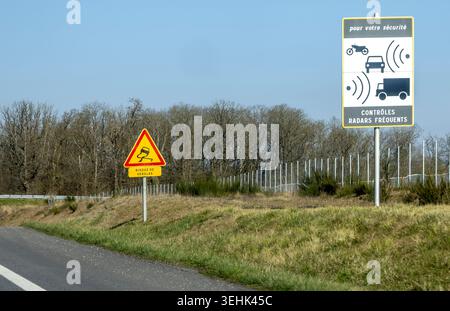 Due cartelli stradali si trovano su un vergine erboso accanto a una strada asfaltata, con una linea di alberi senza foglie e una recinzione sullo sfondo. Foto Stock