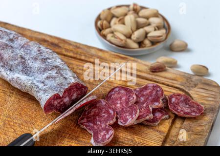 Un coltello affetta una salsiccia stagionata su una tavola di legno accanto a diverse fette. Sullo sfondo sono presenti una ciotola di pistacchi e dadi allentati. Affettare la salsiccia. Bsip 016938 058 Foto Stock