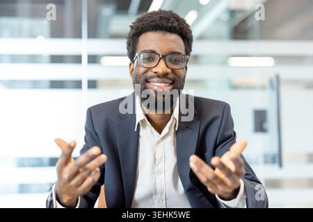 Uomo d'affari sicuro che indossa occhiali da vista e una tuta, seduto in ufficio, comunica e condivide la sua visione mentre gesti attivamente con le mani durante una riunione online Foto Stock