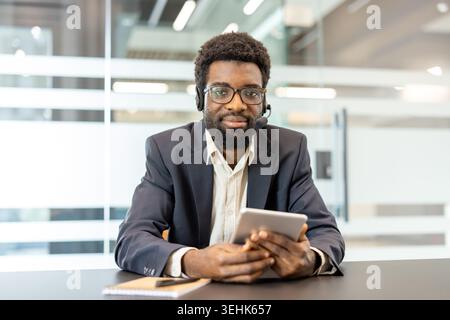 Giovane uomo che indossa cuffie e occhiali, che tiene in mano un tablet digitale e fornisce assistenza clienti online o supporto tecnico da uno spazio di lavoro moderno Foto Stock