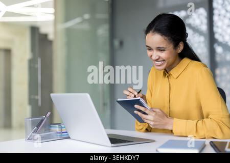Giovane donna d'affari sorridente che lavora dall'ufficio, prende appunti in un notebook mentre guarda un notebook, rappresenta l'istruzione, il lavoro da remoto, la formazione e i concetti di e-learning Foto Stock