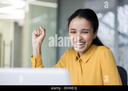 Giovane professionista entusiasta che festeggia alla sua moderna scrivania con un pugno rialzato e un ampio sorriso, esprimendo trionfo, successo e gioioso successo mentre lavora su un notebook Foto Stock