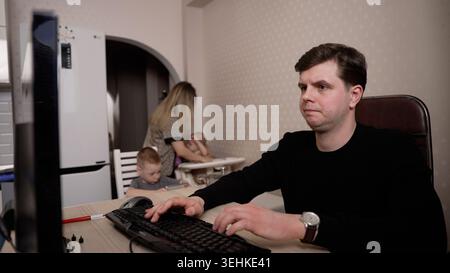 Un giovane stressato lavora al computer a casa, sentendosi frustrato dalle distrazioni della sua famiglia sullo sfondo. Una rappresentazione del lavoro a distanza c Foto Stock