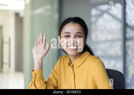 Giovane donna professionale in un ufficio luminoso che sorride e ondeggia alla fotocamera, trasmettendo un calore amichevole e sicuro e una comunicazione positiva in un ambiente aziendale moderno Foto Stock