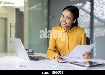 Giovane donna d'affari sorridente con fiducia alla sua scrivania, rivedendo documenti e prendendo appunti con notebook e tablet nelle vicinanze, proiettando professionalità, produttività e leadership Foto Stock