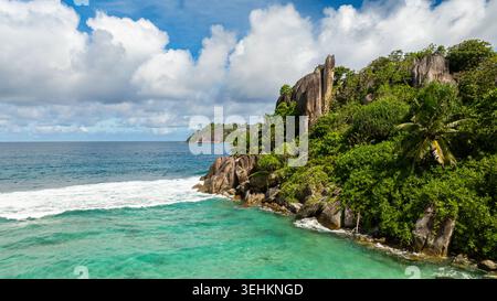 Acque turchesi che incontrano scogliere rocciose circondate da una fitta e lussureggiante vegetazione sotto un cielo luminoso. Seychelles, Mahe. Foto Stock