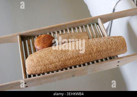 Spugna di loofah naturale, sapone rotondo e una spazzola per unghie in legno appoggiata su un ponte di caddy per vasca da bagno in legno naturale attraverso una vasca bianca. Foto Stock