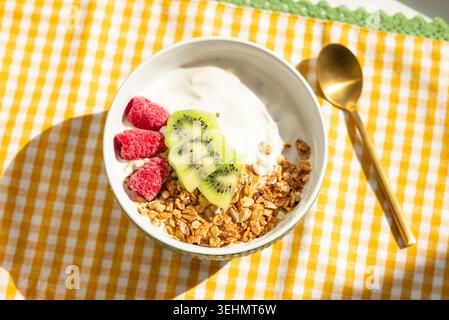 Vista dall'alto di una ciotola per la colazione allo yogurt con farinata d'avena alla muesola, kiwi affettato e lampone congelato su una tovaglia gialla, con un cucchiaio dorato in bri Foto Stock