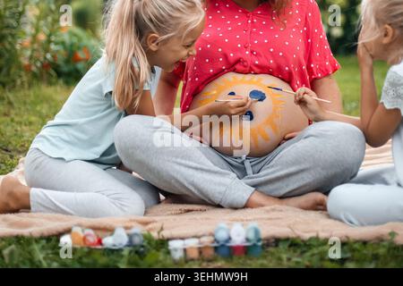 Momenti felici in famiglia all'aperto mentre due bambini dipingono un sole sorridente sulla pancia di una donna incinta. Concetto di gravidanza caldo e giocoso che celebra la maternità, il legame Foto Stock