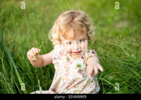 Adorabile bambina bionda con un abito floreale gioca in erba verde alta, raggiungendo un piccolo fiore selvatico in una soleggiata giornata estiva in un prato naturale. Foto Stock