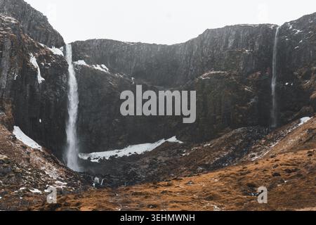 Spettacolare vista della cascata di Grundarfoss che si tuffa lungo le scogliere scure di basalto a Snaefellness, Islanda, con macchie di neve invernali, aria nebbiosa e aspro tumulo Foto Stock