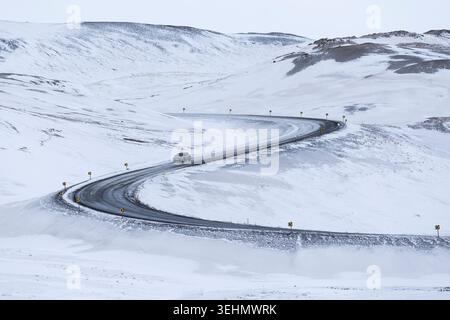 Un'auto solitaria percorre una strada tortuosa attraverso gli altipiani invernali innevati dell'Islanda, con colline bianche e scarse marcature sotto un cielo coperto, evocando re Foto Stock