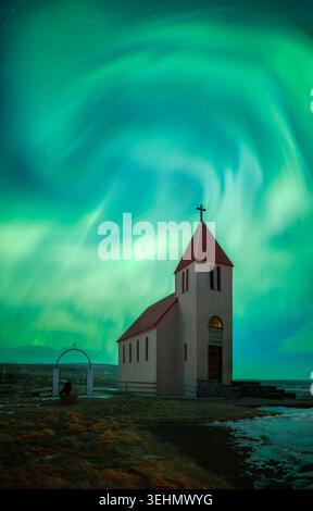 l'aurora boreale verde turbinante si illumina sopra una piccola chiesa islandese con un tetto rosso, situata in un tranquillo paesaggio rurale di notte sotto un cielo stellato. Foto Stock