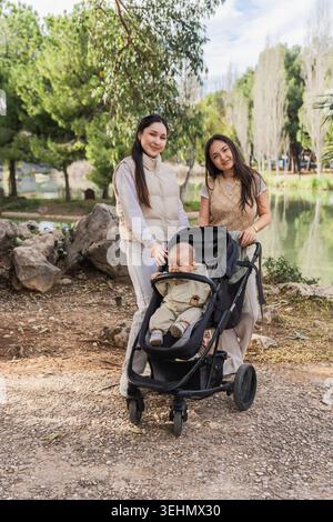 Un momento di gioia in famiglia catturato in un tranquillo parco, con due donne che interagiscono felicemente con un bambino seduto in un passeggino, circondato dalla natura Foto Stock