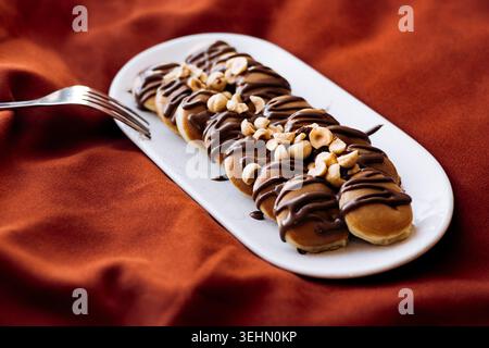 Primo piano di mini frittelle ricoperte da un piatto bianco, ricoperte da un ricco gocciolamento di cioccolato e nocciole tritate, servite come deliziosa colazione o dolce Foto Stock