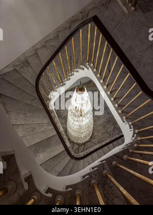 Vista dall'alto di un'elegante scala a chiocciola in marmo con balaustra in ottone, corrimano scuro e un lampadario di cristallo luminoso sottostante, che crea un lussuoso interi Foto Stock