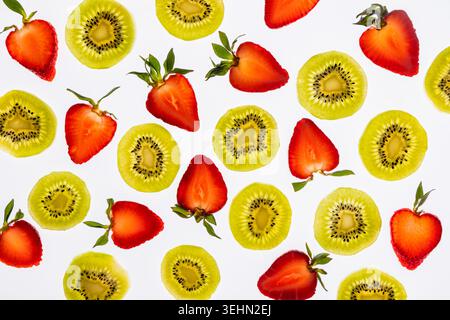 Vista dall'alto, distesa piatta di fette di kiwi fresche e fragole dimezzate sparse su uno sfondo bianco, creando un colorato motivo di frutta estiva per il cibo e. Foto Stock