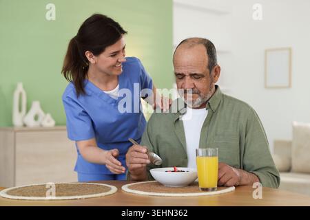 L'accompagnatore fa colazione a un anziano al tavolo di legno all'interno Foto Stock