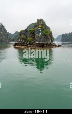 Una piccola isola rocciosa con un faro e vegetazione sorge dalle acque color smeraldo della baia di ha Long, in Vietnam Foto Stock