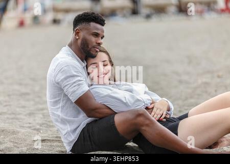 In un pomeriggio di sole alla spiaggia di Zapillo ad Almeria, in Spagna, una coppia condivide un momento tenero rilassandosi sulla sabbia. Si abbracciano comodamente, immergendosi Foto Stock