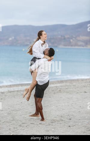 Una coppia gioiosa si impegna in un abbraccio giocoso sulle rive sabbiose di Playa del Zapillo, Almeria. La donna viene sollevata dal suo partner mentre condividono la risata Foto Stock