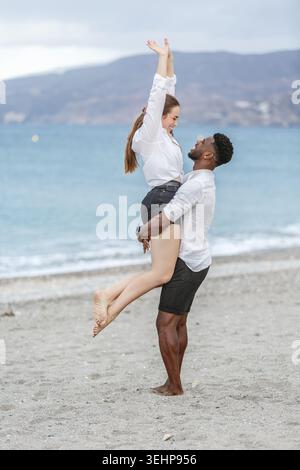 Una coppia condivide un momento delizioso sulla spiaggia sabbiosa, con l'uomo che solleva la donna giocosamente. Sono vestiti in modo informale, trasudando felicità come loro Foto Stock
