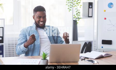 Imprenditore nero guardando laptop, clenching pugni, celebrando il successo aziendale Foto Stock