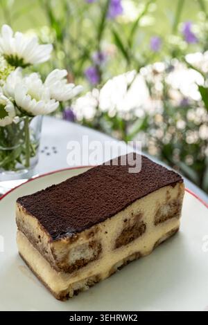 Fetta di tiramisù da vicino con spolverata di cacao in polvere e cremosi strati di mascarpone presso il Garden Cafe all'aperto con fiori di margherita bianchi Foto Stock