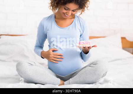 Giovane donna incinta di mangiare una gustosa ciambella, seduta sul letto Foto Stock