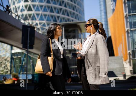 Due donne alla moda si impegnano in una conversazione vivace sullo sfondo di un'architettura moderna sotto un cielo azzurro. Foto Stock