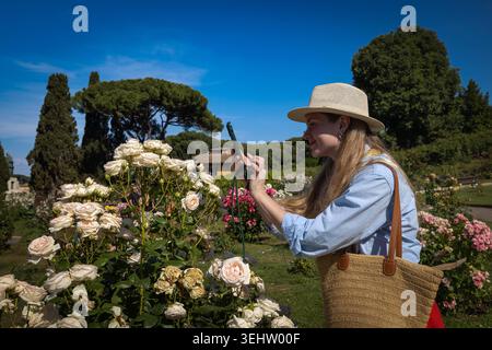 Donna che scatta una foto di rose in fiore al Giardino Civico delle Rose di Roma. Roma, Italia - 24 maggio 2026: Una giovane donna che indossa un cappello di paglia e abiti colorati utilizza il suo smartphone per scattare una foto di un cespuglio di rose bianche in piena fioritura al Giardino delle Rose Municipali di Roma. Lo sfondo mostra il parco pubblico con alberi e altri visitatori sotto un cielo azzurro. Roma RM Italia Copyright: XGennaroxLeonardix Foto Stock