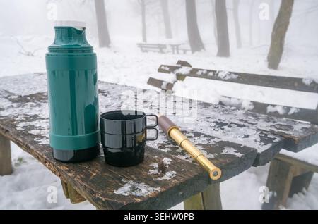 Un thermos verde e una tazza nera sono seduti su un tavolo di legno innevato. La scena è ambientata in un paesaggio invernale con nebbia e panchine innevate nel backgroun Foto Stock