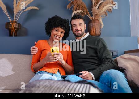 Coppia felice e variegata seduta su un divano, guardando la telecamera. stile di vita domestico. Foto Stock