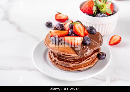 Pila di pancake al cioccolato con una ricca crema di pasta al cioccolato e frutti di bosco freschi - fragole, mirtilli. Colazione dolce e gustosa Foto Stock