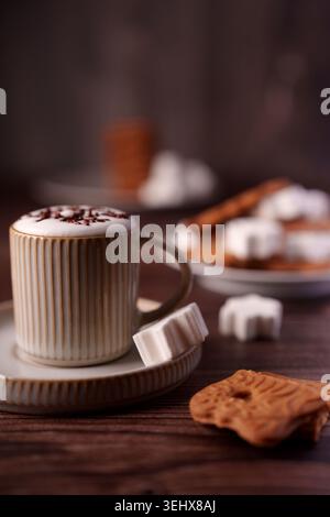 ambiente accogliente di cappuccino con spruzzi di cioccolato in tazza a costine accompagnato da marshmallow sagomati e biscotti su rustici setti da tavola in legno Foto Stock