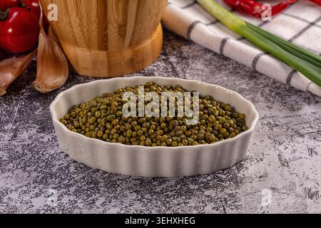 Fagioli verdi mung presentati in un recipiente in ceramica bianca. Cucina rustica con aglio fresco, pomodori rossi, cipolle verdi, peperoncino secco e Malta di legno Foto Stock