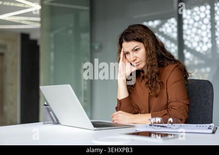 Giovane donna d'affari che si sente sopraffatta e stressata, soffre di mal di testa mentre lavora al suo notebook in un ufficio moderno, simboleggiando l'esaurimento professionale e la pressione sulle scadenze Foto Stock