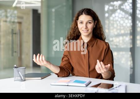 Giovane donna professionista sorridente e gestante alla scrivania durante una videoconferenza, che offre consulenze di lavoro o conduce una riunione online in un moderno ambiente di lavoro Foto Stock