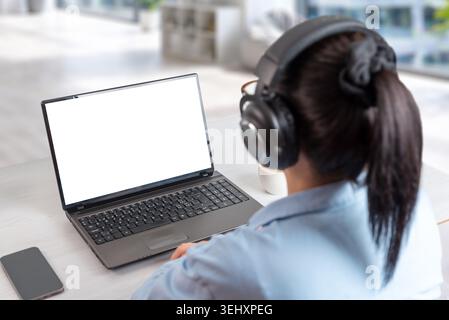 Donna che indossa le cuffie mentre guarda lo schermo vuoto del notebook in uno spazio di lavoro moderno, progettato per l'istruzione online, lo streaming e la comunicazione remota conc Foto Stock
