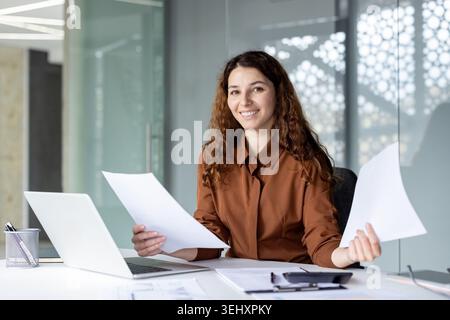 Giovane donna d'affari alla sua scrivania sorride mentre esamina documenti cartacei e report dei notebook in un ufficio moderno e luminoso, trasmettendo produttività, fiducia e attenzione professionale Foto Stock